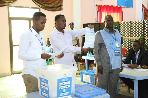 Eine Gruppe von Männern steht um einen Tisch mit Kisten, Papieren und anderen Gegenständen herum und gibt ihre Stimmen bei einer Wahlurne ab, während im Hintergrund Menschen sitzen und stehen.