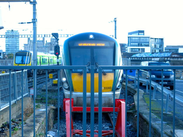 Zwei Züge auf der Schiene mit einem Zaun im Vordergrund, einige Pfosten auf dem Land, einer mit einer Signalleuchte, ein Auto auf der Straße rechts, Gebäude im Hintergrund und der Himmel darüber.