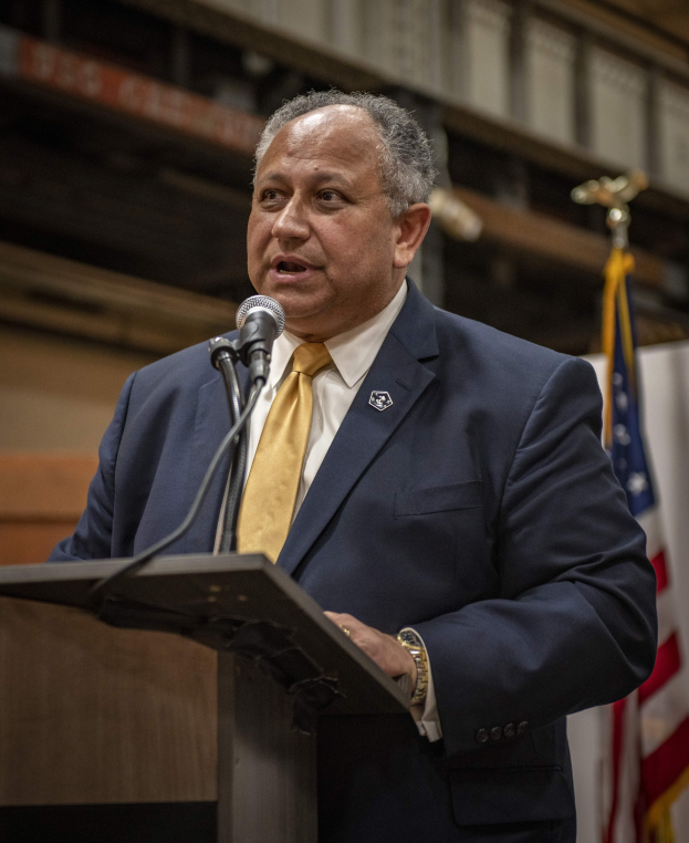 Ein Mann in Anzug und Krawatte steht an einem Rednerpult mit Mikrofon, vor einer Fahne und einer Wand, und scheint eine Ankündigung regarding New York City's neu benannten Bürgermeister zu machen.