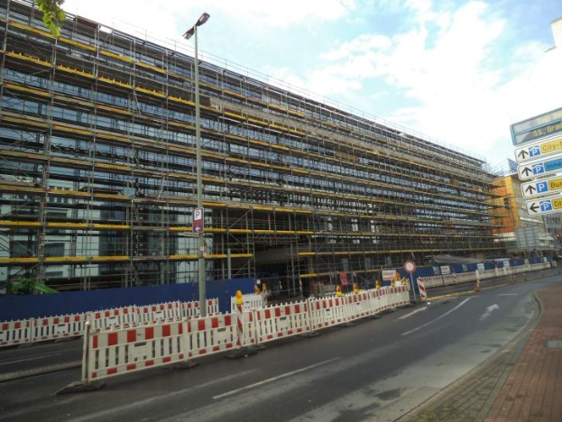 Große Baustelle mit Gerüt, umgeben von Absperrungen, Schildern, Straßenlaternen, Bäumen und einem bewölktem Himmel.