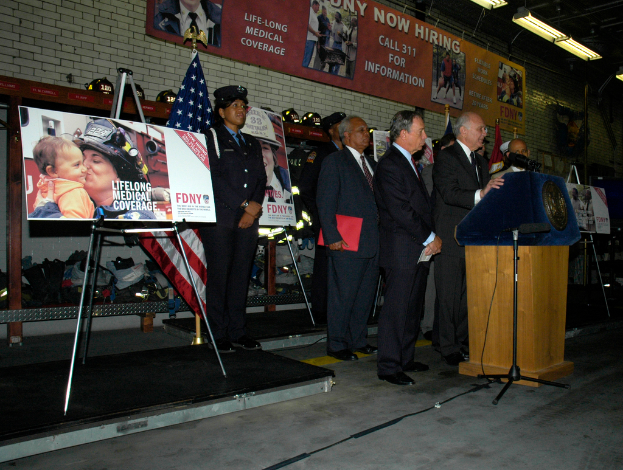 Eine Gruppe von Menschen, die in der Nähe eines Podiums mit Mikrofonen, Schildern auf Ständern, Fahnen und Schildern an der Wand stehen, alles unter hellen Scheinwerfern.
