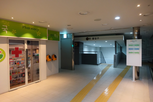 Apotheken-Eingang mit einem "Apotheke"-Schild links, einer Tafel mit Text rechts, beleuchteten Lampen oben und undeutlichen Hintergrundobjekten.