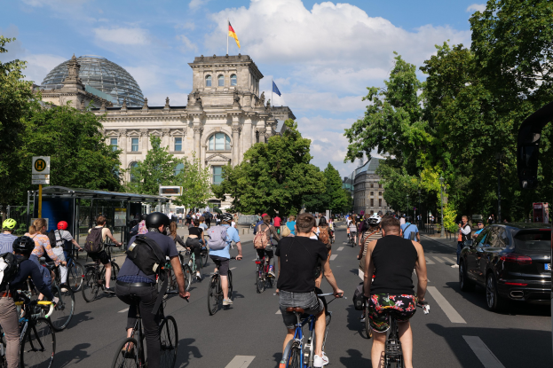 Eine Gruppe von Menschen, die Fahrräder auf einer von Bäumen gesäumten Straße in Berlin, Deutschland, fahren, mit Gebäuden und einer Bushaltestelle im Hintergrund, unter einem bewölkten Himmel und einer Flagge auf einem der Gebäude.