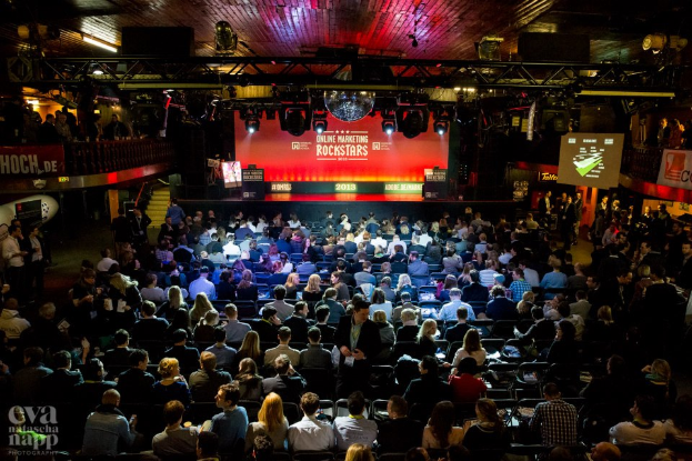 Eine große Menschenmenge sitzt auf Stühlen vor einer Bühne mit Bildschirmen und Bannern, beleuchtet von Scheinwerfern und umgeben von Absperrungen, mit einem Wasserzeichen in der linken unteren Ecke, das eine Atmosphäre der Aufregung und Vorfreude für ein Online-Marketing-Rockstars-Event schafft.