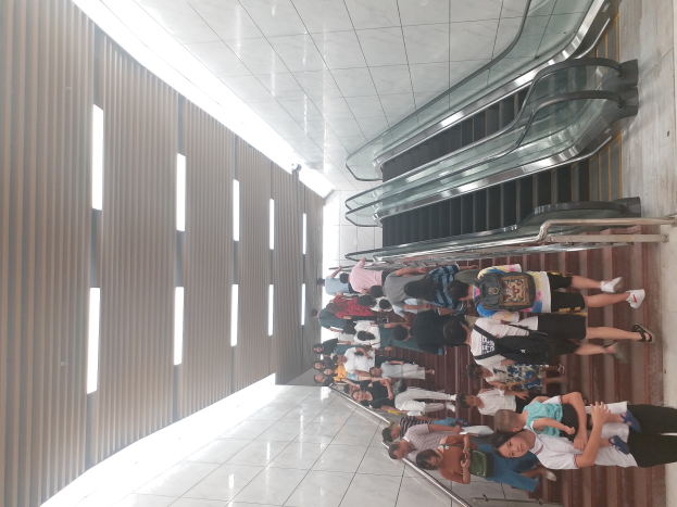 Eine Gruppe von Menschen steht in einer Schlange am Flughafen, trägt Taschen und wartet auf ihren Flug, mit einer Rolltreppe im Hintergrund und sichtbaren Flughafenwänden links.
