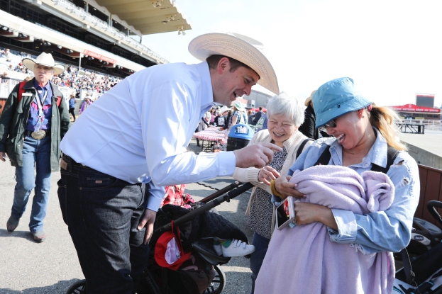 Ein Mann in einem weißen Hemd und Hut hält ein Baby in einem Kinderwagen, umgeben von Menschen in bunten Kleidern und Hüten, mit einer Menge, die auf einem Stadion im Hintergrund sitzt.