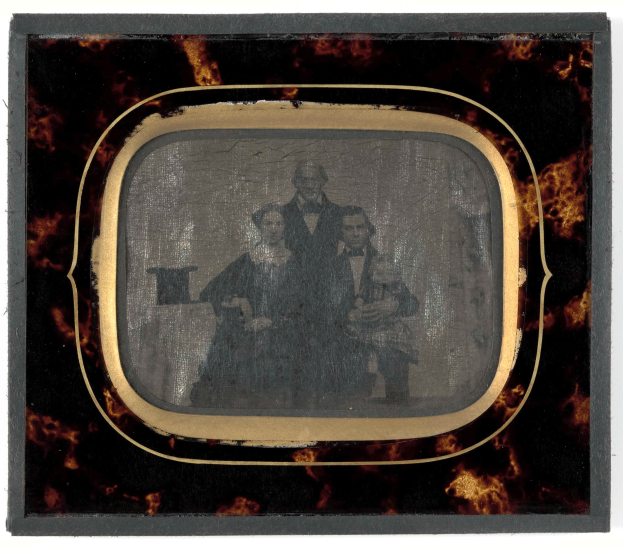 Ein altes Foto eines Mannes im Anzug und Krawatte und einer Frau in unbekannter Kleidung vor einem Fernseher mit ernsten Gesichtern.