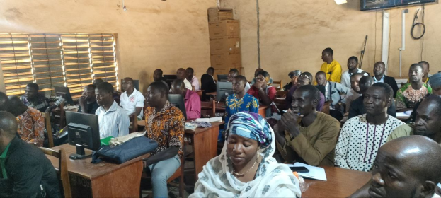 Eine Gruppe von Menschen sitzt an Tischen in einem Klassenzimmer mit Monitoren, Taschen, Büchern und Papieren auf den Tischen, ein Fenster auf der linken Seite und Kartons und einen Fernseher an der Wand im Hintergrund, an einer Techniktrainingssitzung teilnehmend.