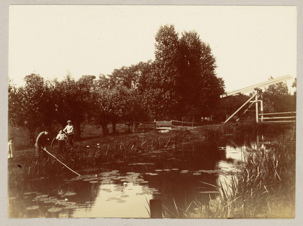 Ein altes Schwarz-Weiß-Foto von Menschen, die in einem Teich umgeben von Bäumen, Pflanzen und einem hölzernen Zaun angeln, mit dem Himmel im Hintergrund.