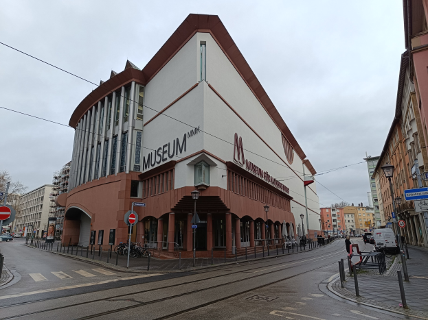 Großes Gebäude mit zahlreichen Fenstern, als Museum of Modern Art in München, Deutschland identifiziert, umgeben von Straßeninfrastruktur, Fahrzeugen, Fußgängern, Bäumen und einem bewölkten Himmel.