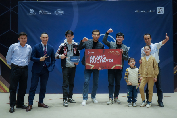 Gruppe von Menschen auf einer Bühne mit einer Tafel mit dem Text "Akang Kuchaydi - Zakovat Quiz", mit einem Banner, einem Barcode und Lichtern im Hintergrund.