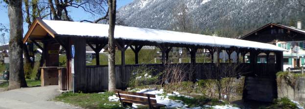 Ein hölzernes Unterschlupf mit schneebedecktem Dach, einer Bank davor, Pflanzen, Gras, einer Gruppe von Bäumen, Häusern mit Dächern und Fenstern, einem Schild, einem Müllcontainer, Pfählen, Hügeln und einem bewölkten Himmel.