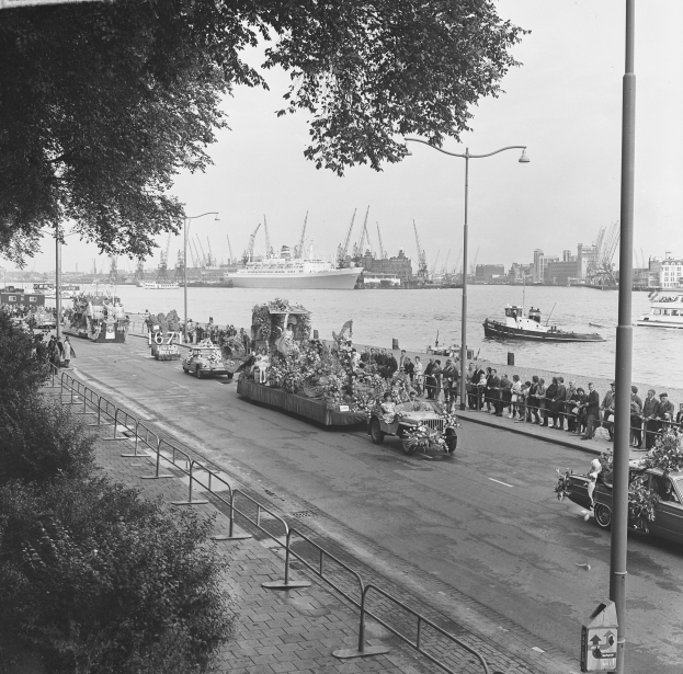 Ein Schwarz-Weiß-Foto eines Umzugs auf einer Stadtstraße, mit Fahrzeugen auf der Straße, Menschen auf dem Gehweg, Geländern, Pfählen, Laternen, Bäumen, Schiffen auf dem Wasser, Gebäuden und einem Himmel im Hintergrund.