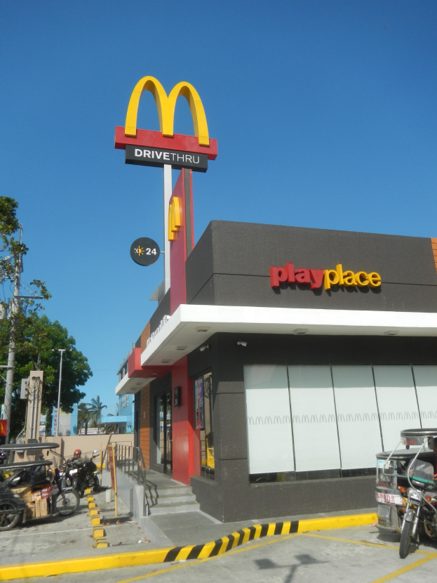 Außenansicht eines McDonald's-Restaurants mit einem "Playplace"-Schild, Fahrzeugen auf der Straße, Bäumen, Strommasten und einem klaren blauen Himmel.