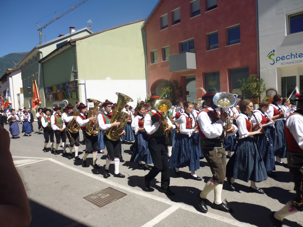 Eine Gruppe von Menschen in traditioneller bayrischer Tracht, die auf der Straße musizieren und dabei durch eine Straße mit Gebäuden gehen, einige halten Fahnen, mit einem Hügel und einem klaren blauen Himmel im Hintergrund.
