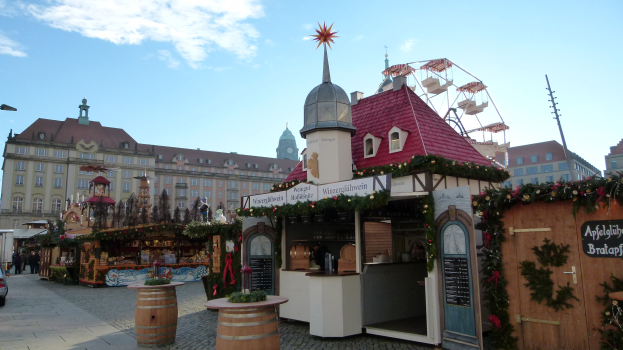 Lebendiger Weihnachtsmarkt in einer Stadtmitte mit geschmützten Bäumen, Gebäuden, Fahrzeugen, Passanten und Wolkenhimmel.