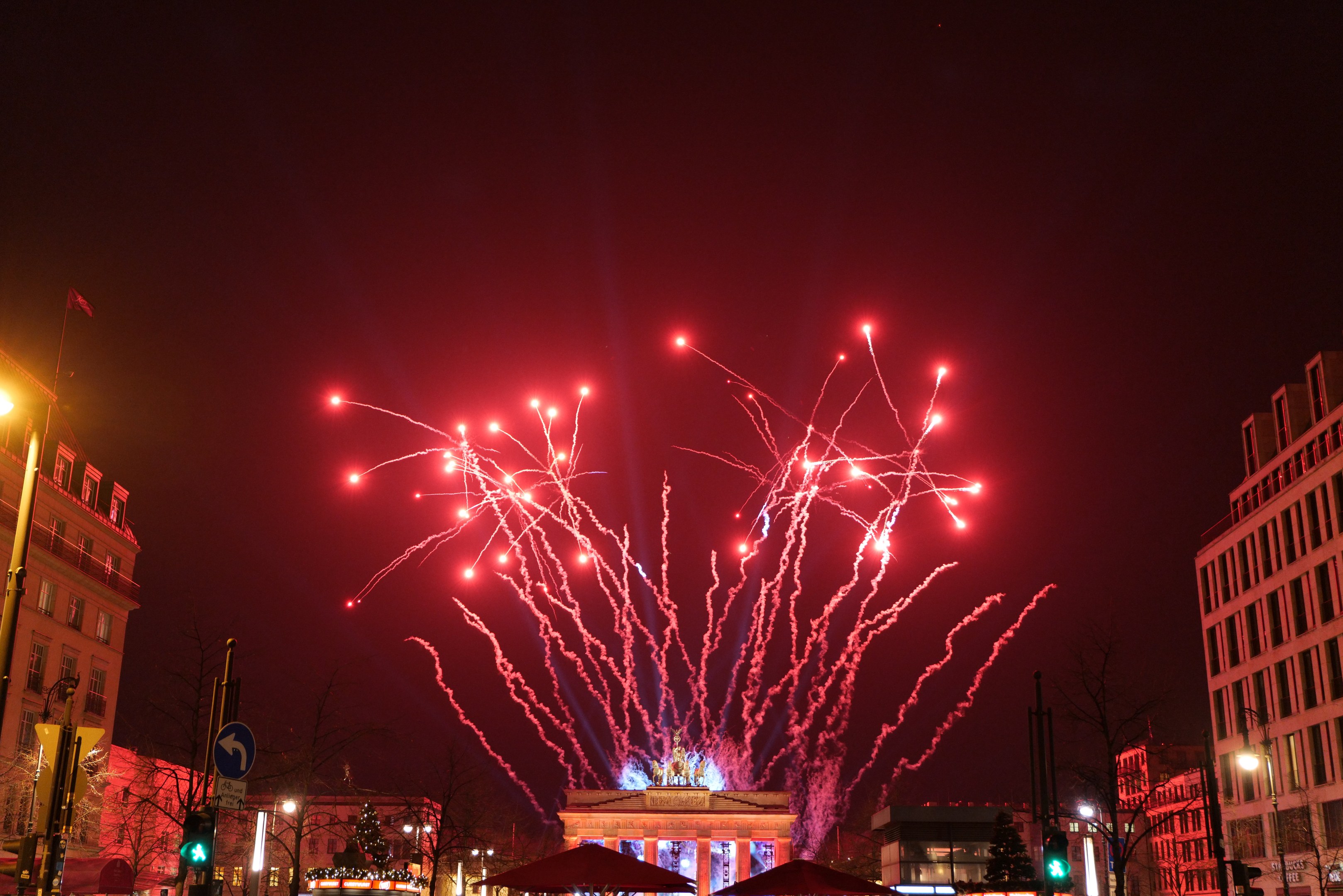 Eine belebte Stadtstraße an einem Neujahrsabend in Berlin, geschmückt mit Gebäuden, Bäumen, Laternen, Verkehrszeichen, Schildern, Zelten, Menschen und einem prächtigen Feuerwerk, das den Himmel erleuchtet.