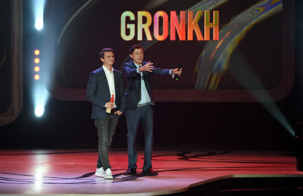 Eine Person in der Mitte hält ein Mikrofon und spricht, während eine andere Person links steht und lächelt, alles vor einem Bildschirm mit Text und Lichtern.