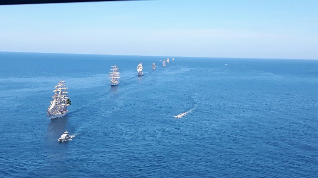 Eine große Flotte von Segelschiffen, die in Formation auf einem klaren blauen Ozean unter einem strahlenden Himmel segeln, mit Sonnenlicht, das sich auf dem Wasser spiegelt.