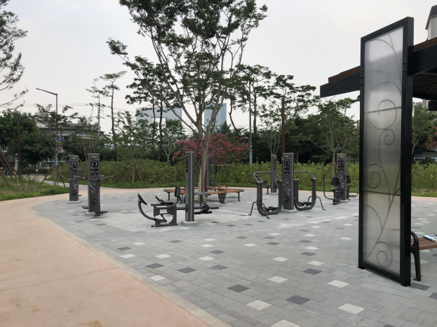 Outdoor park featuring various fitness equipment, benches, trees, plants, grass, poles, lights, wires, and surrounding buildings under a sky backdrop.