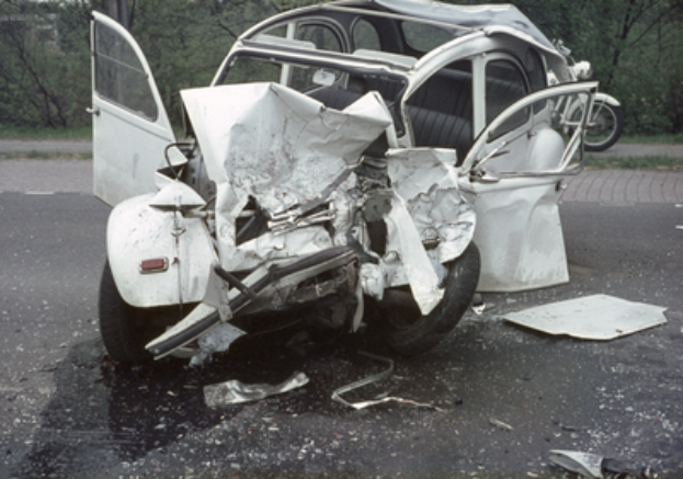 Ein stark beschädigtes Auto mit schwerem Frontschaden steht am Straßenrand, mit Bäumen, einem Gebäude und einem weiteren Fahrzeug im Hintergrund.