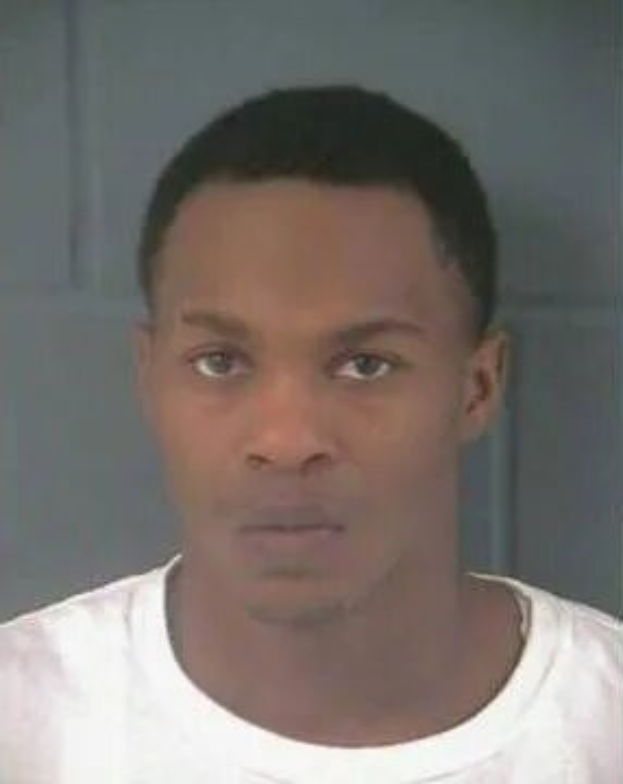 Ein Bild eines Mannes in einem weißen T-Shirt vor einer Wand, der als Verdächtiger in einem Mordfall gilt.