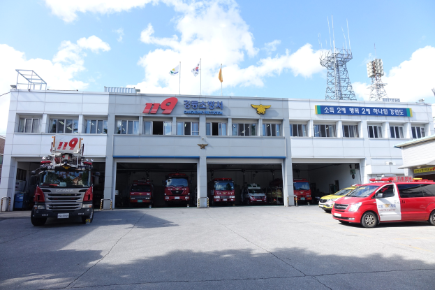 Feuerwache mit mehreren Feuerwehrautos vor dem Gebäude geparkt, Gebäude mit Fenstern und Text, Flaggen mit Stangen im Hintergrund, bewölkter Himmel und fernes Turm.