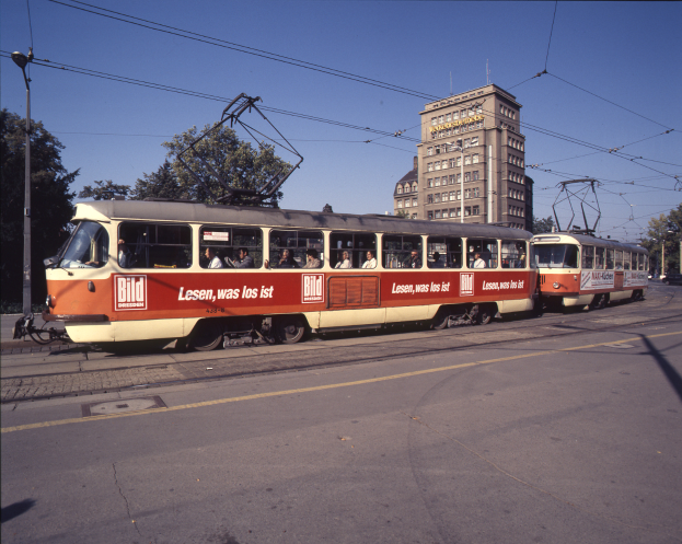 Eine rote und weiße Straßenbahn auf einer Stadtstraße mit Menschen drinnen, gesäumt von Bäumen, Pfählen und Drähten, mit Gebäuden und einem klaren blauen Himmel im Hintergrund.