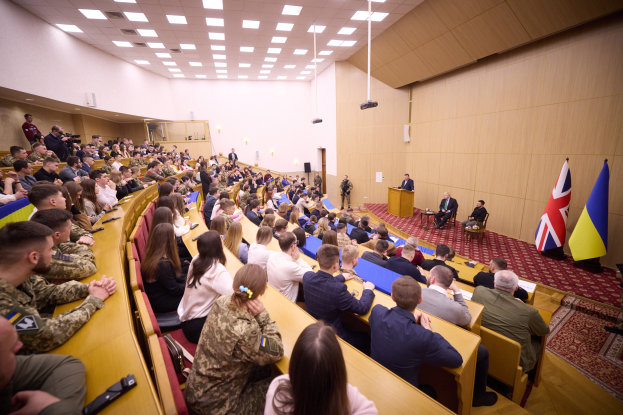 Eine große Gruppe von Menschen sitzt an Tischen in einem Raum mit Flaggen auf der rechten Seite, einem Podium mit Mikrofon vorne und einem Projektor und Lampen an der Decke, was auf eine Militärakademie hindeutet.