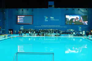 Gruppe von Menschen um ein Hallenbad mit Netz am Boden herumstehend, vor einer Wand mit "FINA World Championships 2015 Kazan" auf einem Bildschirm, mit Trägern und Lampen darüber.