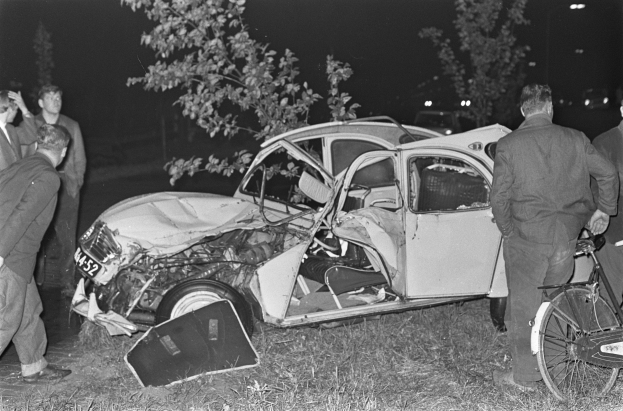 Ein Schwarz-Weiß-Bild eines auf der Straße liegenden Wagens mit mehreren Menschen drumherum, einer hält ein Fahrrad, und Bäume mit Lichtern im Hintergrund.