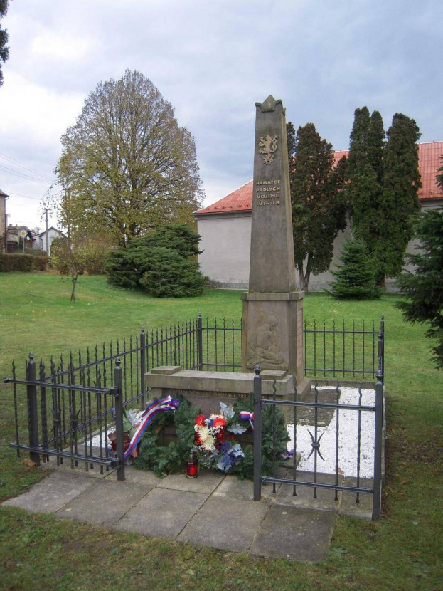 Ein Denkmal auf einer Wiese mit einem Kranz, umgeben von einem Metallzaun, das Holocaust-Opfern in Vostiu, Polen gedenkt, mit Bäumen, Gebäuden und einem klaren blauen Himmel im Hintergrund.