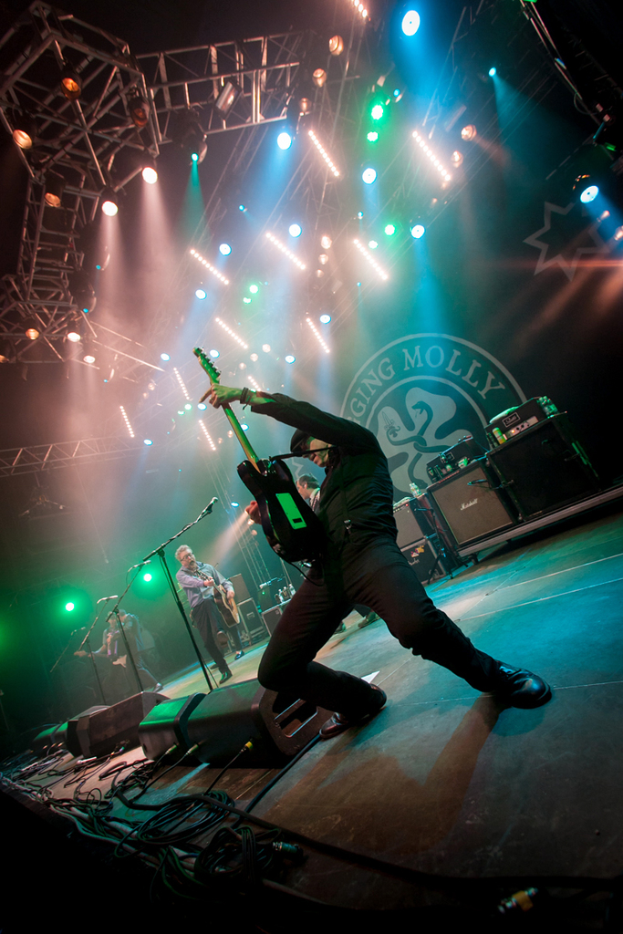 Ein musikalisches Konzert mit drei Performern auf der Bühne: zwei Gitarristen rechts mit Mikrofonen davor, ein Hintergrund mit dem Wort "Molly" und zwei Schlangensymbolen sowie Scheinwerfer darüber.