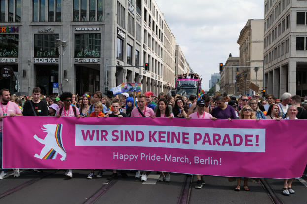 Eine Gruppe von Menschen marschiert auf einer Straße in Berlin, Deutschland, mit einer pinken "Happy Pride March"-Fahne, während Gebäude, Laternenpfähle und Verkehrszeichen die Straße säumen und der Himmel bewölkt ist.