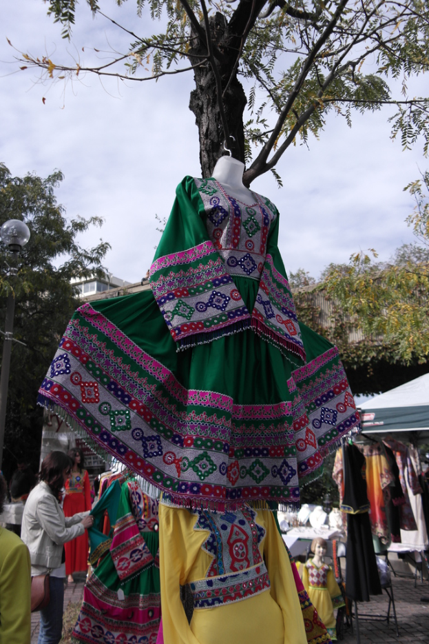 Eine Schaufensterpuppe steht inmitten eines belebten Einkaufsgebiets mit verschiedenen Geschäften im Hintergrund, einer Person, die Kleider an einem Pfahl arrangiert, einem Gebäude und Bäumen daneben.