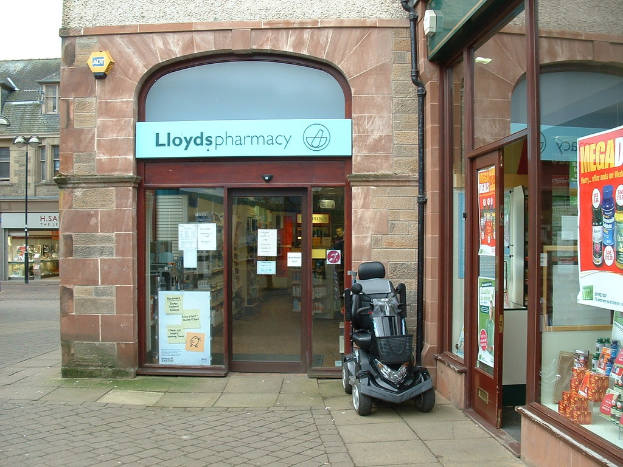 Eine Apotheke mit einem vor ihr geparkten Fahrzeug und einem Gebäude in der linken Ecke.