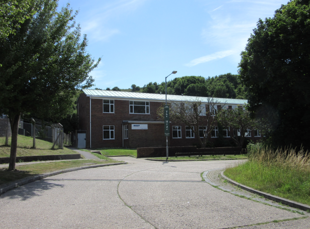 Ein Schulgebäude mit Fenstern, einem Straßenmast, einem Schild, einer Baumgruppe, Gras, einem Zaun und einem bewölkten Himmel, das sich in einem Park mit saftigem Grün befindet.