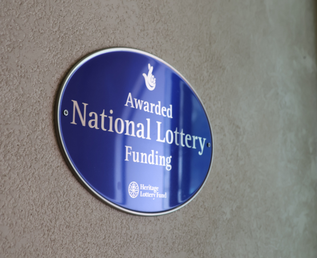 Blaues rechteckiges Schild an einem Gebäude mit weißem Hintergrund und schwarzer fetter Schrift, die "Mit Nationaler Lotterie-Förderung ausgezeichnet" lautet.