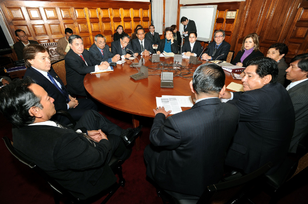 Eine Runde Tischbesprechung mit Delegierten, die diskutieren, Mikrofone, Papiere, Kabel und Dateien auf dem Tisch, mit einer hölzernen Wand im Hintergrund.