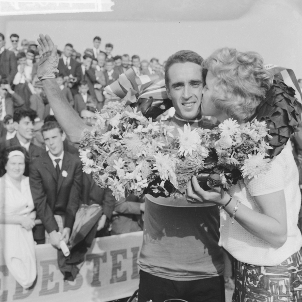 Ein Schwarz-Weiß-Foto eines Mannes und einer Frau, die sich in einer Menschenmenge umarmen und küssen, wobei der Mann einen Blumenstrauß hält und die Frau strahlt, während die Menge applaudiert.