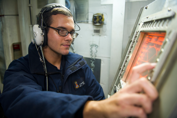 Ein Mann in einer blauen Uniform, mit Kopfhörern und Brille, arbeitet konzentriert an einem Computer mit verschiedenen Gegenständen im Hintergrund.