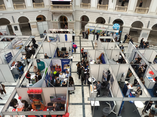 Große Gruppe von Menschen auf einer internationalen Fachmesse mit Ständen, Bannern, Tischen, Stühlen, Ballons und einem Gebäude mit Bögen, Säulen, Geländern und einem Bildschirm im Hintergrund.