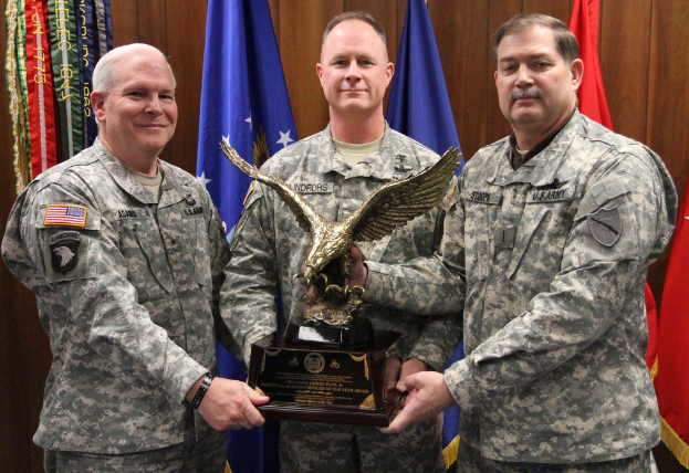 Drei Männer in Uniformen stehen vorn, jeder hält eine Auszeichnung, mit Fahnen dahinter und bunten Dingen mit Text oben links.