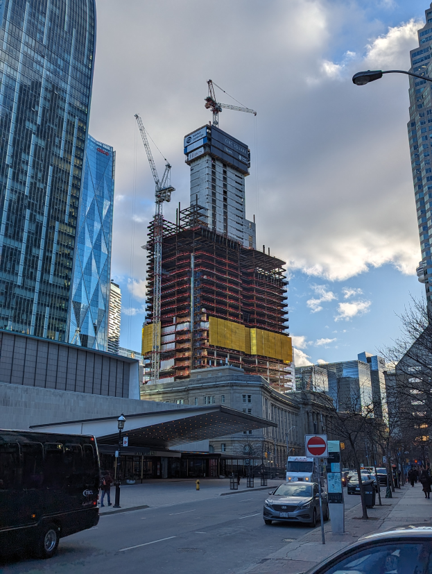 Eine belebte Stadtstraße mit Fahrzeugen, Fußgängern, Schildern, Laternen, Bäumen und dem One World Trade Center im Hintergrund unter einem bewölkten Himmel.