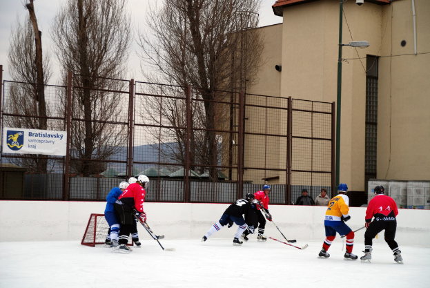 Menschen, die Eis-hoc-key auf einem Eisstadion mit Gebäuden, Bäumen, einer Straßenlaterne, einem Namensschild und Zäunen im Hintergrund unter einem Himmel spielen.