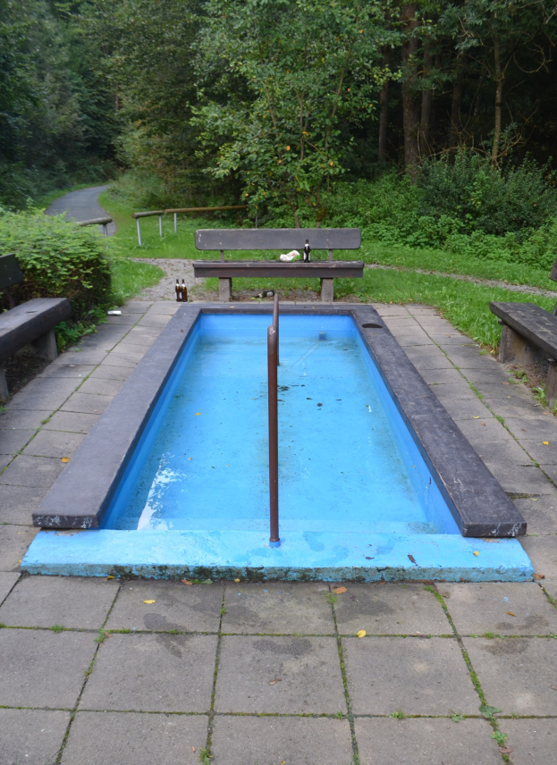 Kleiner Parkteich mit zentralem Pfahl, umgeben von Bänken, Grün, Straße und Bäumen.