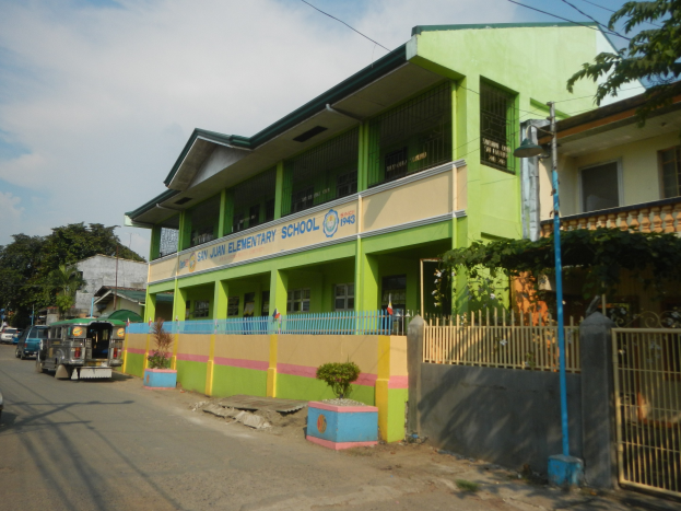 Schulgebäude mit grüner und gelber Fassade neben einer Straße, umgeben von Fahrzeugen, Blumentöpfen, einem Zaun, einem Tor, einem Straßenpfahl, Bäumen, Drähten und einem bewölkten Himmel.