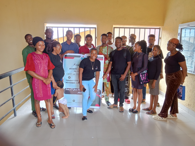 Eine Gruppe von Menschen, die in einem Raum zusammenstehen und ein Schild in der Mitte halten, mit einem Geländer auf der linken Seite, Fenstern und einer Wand im Hintergrund.