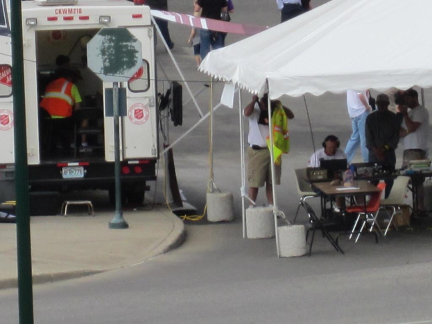 Ein Rettungswagen steht auf der linken Seite einer Stra├če, w├Ąhrend auf der rechten Seite ein Zelt viele Menschen sch├╝tzt, mit einem Tisch und einem Stuhl in der N├Ąhe.