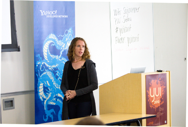 Eine Frau in einem schwarzen Kleid steht neben einem Rednerpult in einem Konferenzraum, mit einem Tisch neben dem Rednerpult und einem Banner, einer Wand und einem Fenster im Hintergrund.